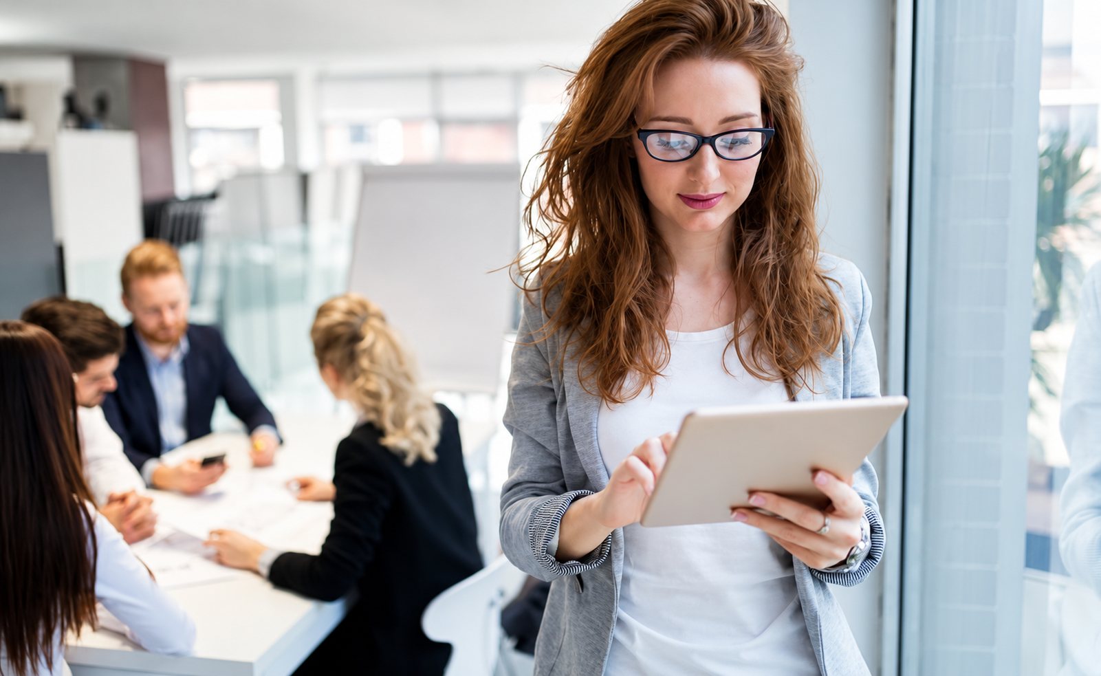 Une femme aux cheveux roux et à lunettes consulte une tablette au premier plan, tandis que des collègues sont réunis autour d'une table en arrière-plan.