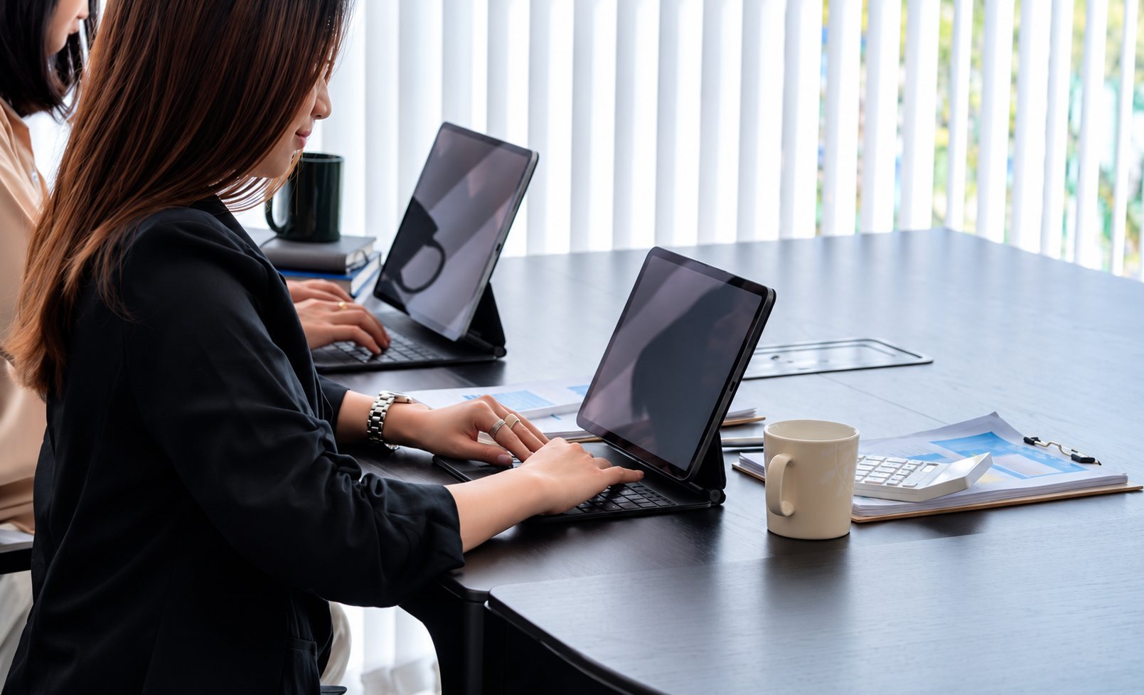 Deux femmes sont assises à une table sombre et tapent sur des tablettes munies de claviers, avec une tasse, une calculatrice et des documents à côté.