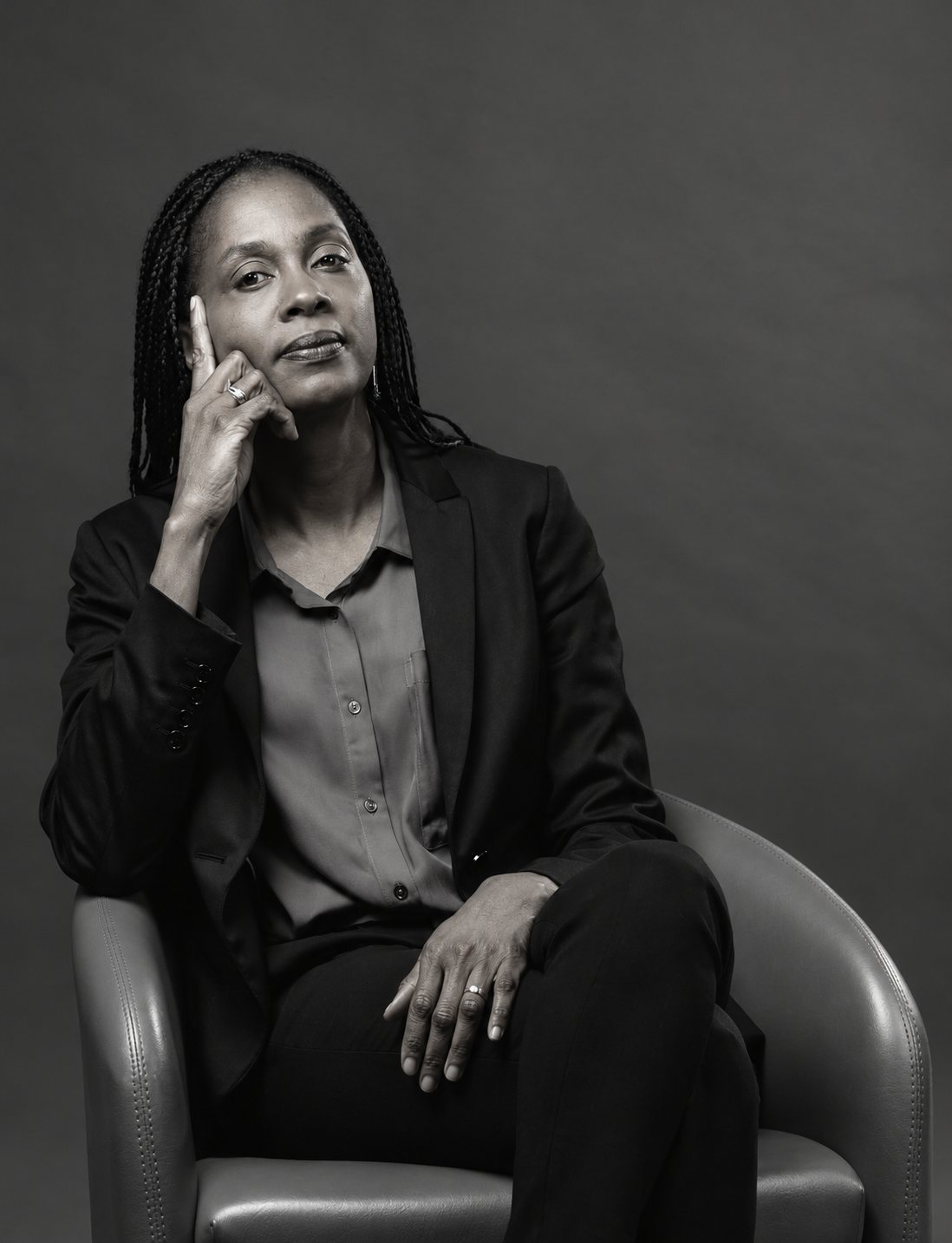 Portrait en noir et blanc d'une femme aux tresses, vêtue d'une veste sombre, assise dans un fauteuil en cuir et la main contre la tempe.