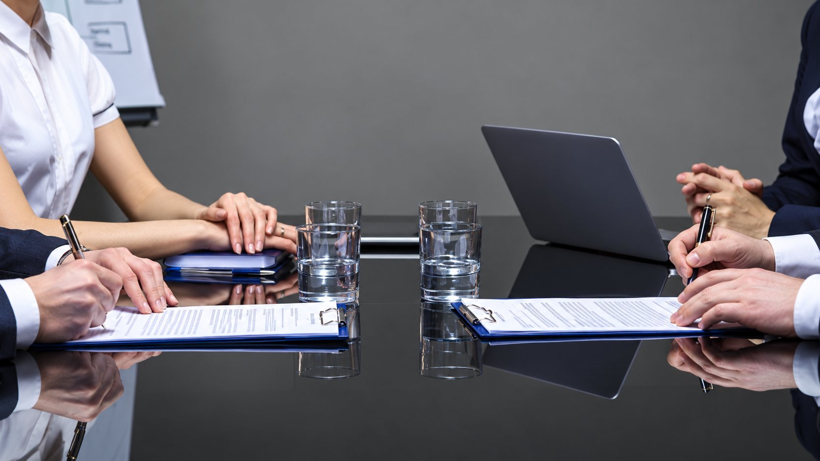 Des personnes en tenue professionnelle sont assises autour d'une table vitrée sur laquelle sont posés des documents, un ordinateur portable et des verres d'eau.